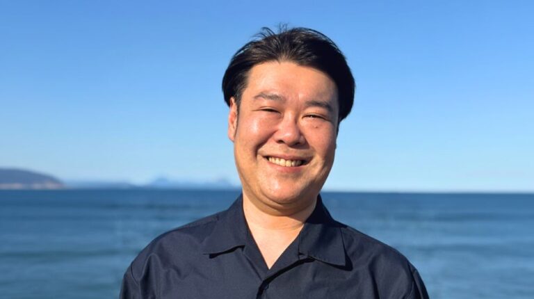 海を背景に笑顔で立つ工藤悠平氏のポートレート写真