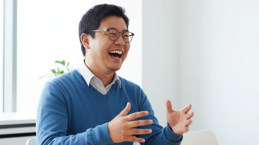 明るいオフィスで笑顔を見せながら話す男性。青いセーターに白いシャツを着て、両手を広げて会話をしている様子。インタビューや対話の場面をイメージさせる写真。